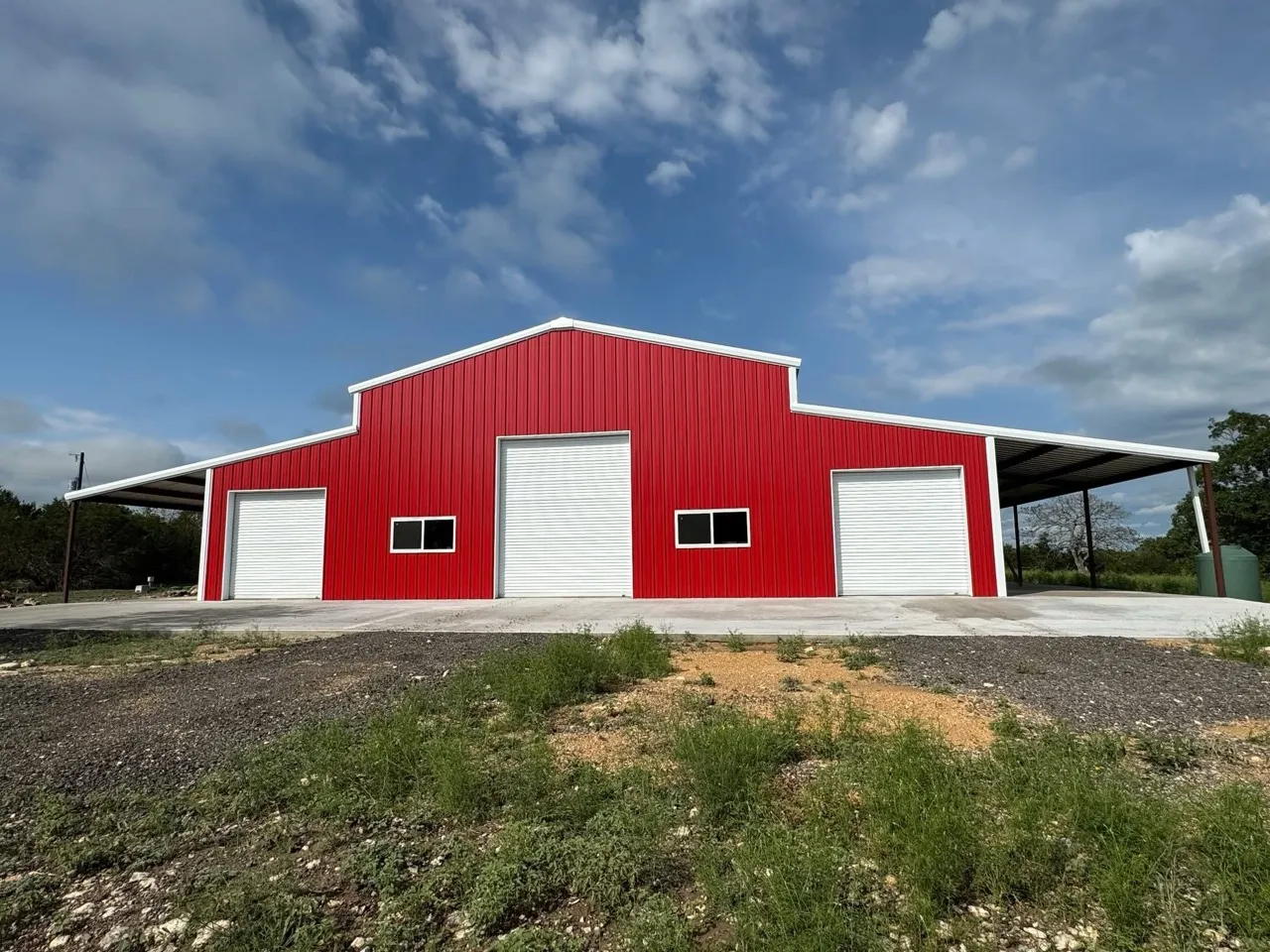 A red metal barn with three white roll-up doors and covered wings on both sides.